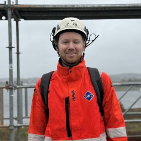 En mann står på en byggeplass og smiler mot kameraet. Han har på seg hvit vernehjelm, en oransje arbeidsjakke med refleks og en svart ryggsekk. I bakgrunnen er det stillaser og overskyet vær.