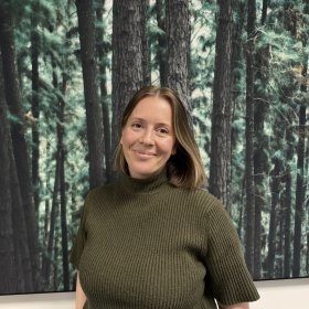 En kvinne med kort, lyst brunt hår står foran et stort bilde av en tett furuskog. Hun smiler svakt og ser rett mot kamera. Hun har på seg en olivengrønn, ribbestrikket topp med høy hals og korte ermer.