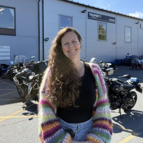 En kvinne med langt, brunt, krøllete hår står utendørs foran en bygning med motorsykler parkert utenfor. Hun smiler mot kamera. Hun har på seg svarte klær og en fargerik, stripete cardigan. Det er solskinn og blå himmel.