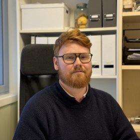 En mann med briller og rødt hår og skjegg sitter i en kontorstol på et kontor. Han har på seg en mørk strikkegenser og ser rett mot kamera. Bak ham står hyller med permer, esker og kontorutstyr.