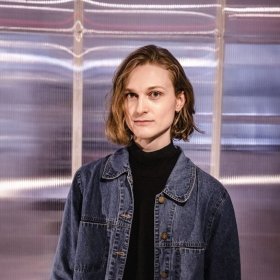 En person med skulderlangt, bølget hår ser rett mot kamera. Hen har på seg en mørk høyhalset genser og en dongerijakke. Bakgrunnen består av et transparent, bølget materiale med lysrefleksjoner i blått og rosa.
