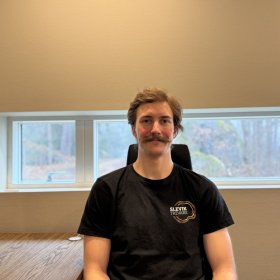 En mann sitter ved siden av et skrivebord og ser rett mot kamera. Han har bart og kort, brunt hår og har på seg en svart t-skjorte med logoen «Slevik Trevare». Bak ham er et vindu med utsikt mot skog.