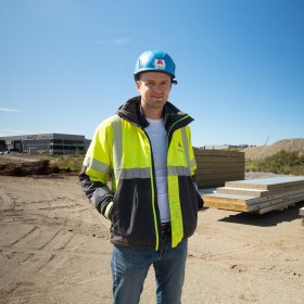 En mann står med hendene i lommene og smiler til kamera. Han har på seg hjelm og refleksjakke, og står på et stort åpent område med byggematerialer i bakgrunnen.