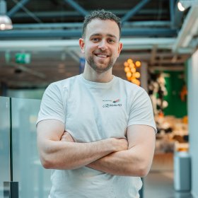 En mann i hvitt arbeidstøy står med armene i kors og smiler til kamera.