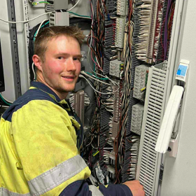 Maritim elektriker Arne Johan Svendsby foran brann og gass-kabinettet Mann med lyst hår sitter foran kabinett med mange ledninger. Han har gul arbeidsjakke på.