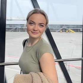 Lettmatros Hedda Othilie Hildre Dame med lysegrønn t-skjorte og lyst hår i hestehale står i en gang med glassvegger på en flyplass