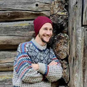 Mann med tradisjonell ullgsenser og rød lue lener seg mot en vegg av tømmer