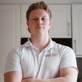 En ung mann med lyst hår og hvit poloskjorte står med armene i kors og smiler til kamera.