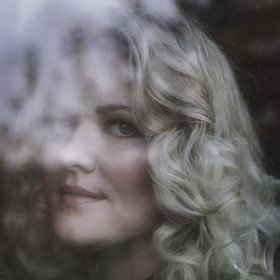 Et nærbilde av ansiktet til en kvinne med lyst, bølgete hår. Hun ser inn i kamera gjennom en dugg- eller regnvåt rute, noe som gjør deler av ansiktet uklart.