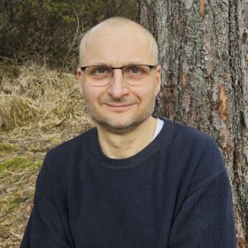 Mann med briller og svart genser står foran et tre. I bakgrunnen er det skog.