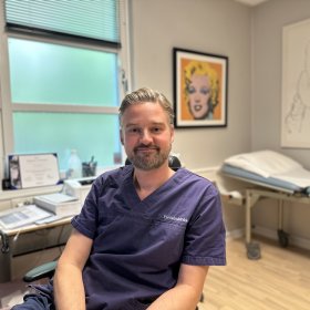 En mann i mørkeblå legeuniform sitter i et lyst legekontor og smiler rolig mot kamera. Bak ham sees en undersøkelsesbenk, medisinsk utstyr og et fargerikt portrett av Marilyn Monroe på veggen.