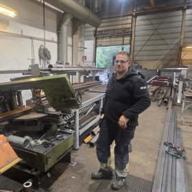 En mann står inne i et verksted omgitt av metallplater, rør og store maskiner. Han har på seg vernebriller, svart arbeidsjakke og mørke arbeidsbukser med refleksdetaljer. I bakgrunnen er det åpne porter som slipper inn dagslys og viser trær utenfor. Verkstedet er fullt av industrielt utstyr, og gulvet er støvete etter arbeid.