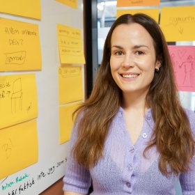 Kvinne med langt, brunt hår i lilla cardigan. Hun ser rett i kameraet og smiler. I bakgrunnen er en tavle og en glassdør dekket med gule og noen rosa post-it-lapper.