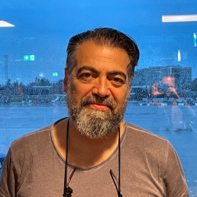 Mann med mørkt hår og skjegg står foran et vindu. Han smiler til kameraet.