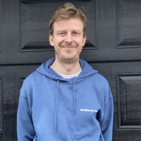 Mann med blå hettegenser står foran en svart dør og smiler.