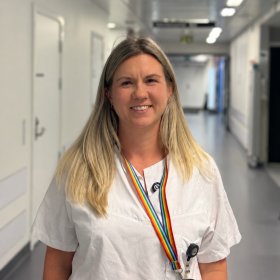 En kvinne med langt, lyst hår står i en korridor på et sykehus og smiler mot kameraet. Hun har på seg hvitt arbeidstøy og et regnbuefarget nøkkelbånd rundt halsen. I bakgrunnen er en lys sykehuskorridor med dører på begge sider.