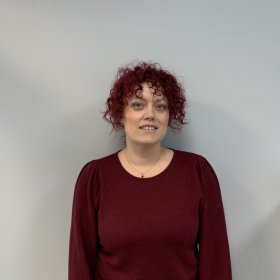 En kvinne med kort, krøllete rødt hår står foran en lys, nøytral vegg og ser rett mot kamera. Hun har på seg en mørk rød genser med lange ermer og et enkelt smykke rundt halsen. Ansiktsuttrykket er rolig og vennlig med et svakt smil.