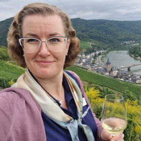 Lyshåret dame med briller står foran en landsby i bakgrunnen som ligger på hver side av en elv. Det er grønt og frodig i landskapet.