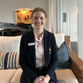 En ung kvinne i mørkeblå uniform sitter i en sofa. Hun har lyserosa skjorte på seg. Hun smiler mot kameraet.