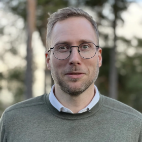 En mann står utendørs og ser rett mot kamera. Han har kort, lyst hår, kort skjegg og runde briller. Han har på seg en grå genser over en hvit skjorte. Bakgrunnen er litt uskarp med trær i dempet lys.