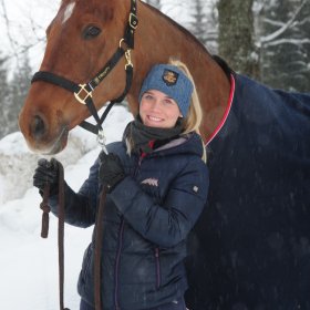 Lyshåret dame med mørkeblå jakke og bukse og pannebånd står foran en brun hest med dekken. Det er noen trær i bakgrunnen. Det er vinter og snø.