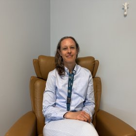 En kvinne i lyse klær med brunt, halvlangt hår sitter i en brun lenestol på et kontor eller møterom. Hun ser inn i kameraet og smiler svakt. Veggene er lyse.