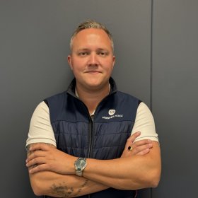 En mann står med armene i kors foran en mørkegrå vegg og ser mot kameraet. Han har på seg en hvit T-skjorte og en mørkeblå vest med Mopedbil Norge-logo. Han har klokke på håndleddet og tatoveringer på armene.