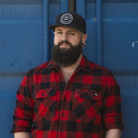 Portefølje- og reiseeffektmaker og salmaker Paul Alexander Vink Mann med skjegg og caps står foran en mørkeblå dør. Han har hendene i lomma, en ring i det ene øret og en rødrutete skjorte.