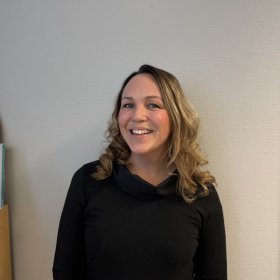 En kvinne med skulderlangt, bølgete hår står foran en lys vegg og smiler mot kamera. Hun har på seg en svart topp med høy hals og lange ermer.