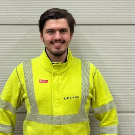 Mann med brunt hår og gul arbeidsjakke med refleks står foran en lys vegg og smiler til kamera