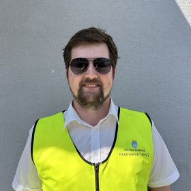 En mann med brunt hår og brunt skjegg smiler. Han har på seg solbriller med mørke glass, en gul refleksvest og en hvit t-skjorte med krage under. Han står inntil en grå vegg.