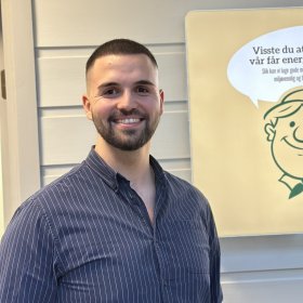 En mann med kort mørkt hår og skjegg står og smiler foran en lys vegg. Han har på seg en mørkeblå skjorte med hvite, tynne striper. Bak ham henger et skilt med en tegnet figur av en smilende mann og teksten: «Visste du at fabrikken vår får energi fra solen? Slik kan vi lage gode matvarer på en enda mer miljøvennlig og bærekraftig måte.»