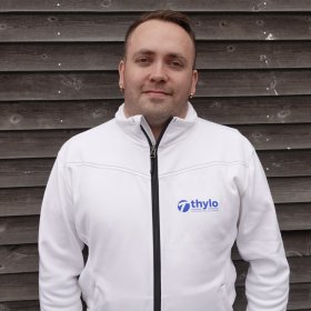 Mann som smiler til kamera med hvit arbeidsjakke med blå logo.