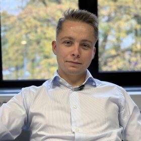 En ung mann med kort, lyst hår og lyseblå, stripete skjorte sitter foran et stort vindu og ser mot kameraet. Utenfor vinduet skimtes trær med høstfarger.