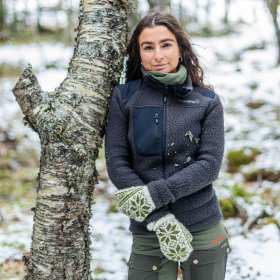 Damed med svart hår og svart fleecejakke og strikkevotter lener seg mot et tre i en skog med litt snø på bakken