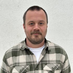 En mann står foran en lys murvegg og ser rett mot kameraet. Han har kort hår, skjegg og bart, og smiler svakt. Han har på seg en grønn- og hvitrutet flanellskjorte med en hvit T-skjorte under.