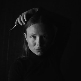 Skomaker Irene Dorthea Johansen Det er et svart-hvitt portrett av en ung kvinne med langt hår som faller bak skuldrene. Hun har et alvorlig uttrykk og ser rett mot kamera. Det ene armen er løftet, og hånden holder en liten metallgjenstand over hodet. Ansiktet er delvis opplyst fra venstre side, mens bakgrunnen og den andre siden av ansiktet er mørk. Hun har på seg en mørk, høyhalset genser.