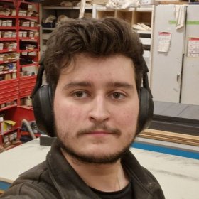 En person står i et verksted og ser rett mot kameraet. Personen har mørkt hår, kort skjegg og bruker store hørselsvern. I bakgrunnen sees hyller med verktøy og materialer, samt arbeidsbord og skap.