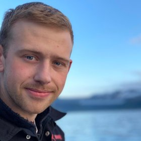 Bildet er et nært portrett av en ung mann som smiler svakt mot kameraet. Han har kort, lyst hår, blå øyne og lett skjeggvekst. Han har på seg en mørk jakke. Bak ham ser man havet og et blålig landskap i myk uskarp bakgrunn, trolig fjell i det fjerne. Himmelen er klar og blå.