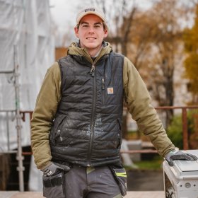 En mann med arbeidsklær og caps står og lener seg på en maskin. I bakgrunnen er det tydelig høst og overskyet. Mannen smiler til kamera.