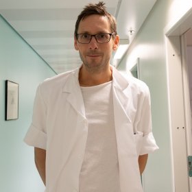 En mann i hvite sykehuseklær og briller. Han ser rett i kameraet. Han er i sykehuset.