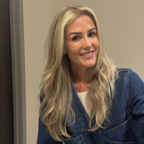 Kvinne med langt blondt hår smiler til kamera. Hun har på seg en mørkeblå dongeri­jakke, med en hvit T-skjorte synlig under.