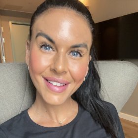 En dame i svart genser smiler til kamera. Hun har blå øyne, brunt hår og håret er i hestehale. Hun sitter i en grå sofa og bak hun er det en svart TV på veggen. 