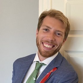 En mann med blondt hår og skjegg smiler bredt. Han har på seg dress - grå dressjakke, hvit skjorte, grønt slips og 17.mai rød sløyfe. Bak han er det en grå vegg og hvit dør.