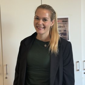 En smilende kvinne med langt, blondt hår i hestehale står foran et hvitt skap. Hun har på seg en mørkegrønn overdel og en svart blazer.
