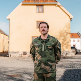 En mann med brunt hår og uniform står med hendene bak ryggen og smiler til kamera. I bakgrunnen er en lys, gammel bygning og blå himmel.