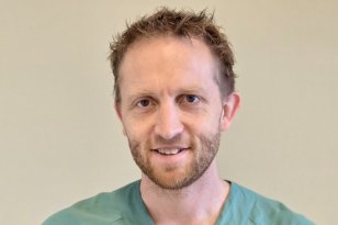 En mann med brunt hår og skjegg smiler. Han har på seg en grønn sykehusskjorte som leger har på seg. På skjorten henger avgangskortet hans, og på høyre side er det en lomme på skjorten hvor det er en penn som stikker opp. Bak mannen er det en beige vegg.