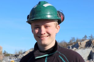 Ung, smilende mann med grønn hjelm og mørk genser. I bakgrunnen skimtes stein, trær og en blå himmel. 