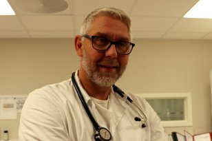 Mann med grått hår og briller har et stetoskop rundt halsen og er kledd i hvitt. 