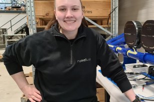 Ung kvinne står på et verksted. Hun er kledd i mørkt arbeidstøy 